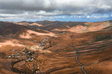 Aerial view of Fuerteventura shows volcanic hills, terraced slopes, whitewashed hamlets, and a winding road toward a valley under low clouds and shifting light. © Aerial Film Studio