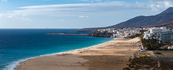 Aerial view of Morro Jable, Fuerteventura, Canary Islands, with golden beach curving to a rocky headland and marina pier, whitewashed hotels, and arid volcanic slopes. © Aerial Film Studio