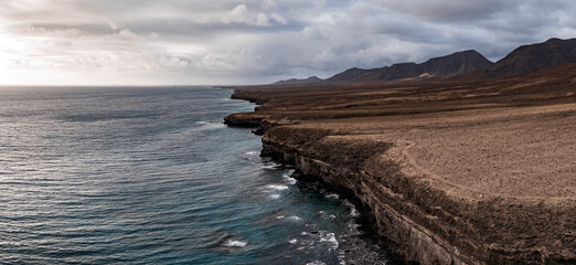 Stratified brown cliffs meet turquoise foaming Atlantic water in Fuerteventura near Playa del Bajo de la Burra, with arid lava plains and conical mountains.