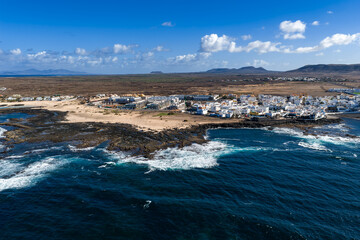 Aerial view of a coastal town on Fuerteventura, Canary Islands. Whitewashed buildings line volcanic rock shelves and sandy coves as Atlantic surf hits jagged lava reefs. © Aerial Film Studio