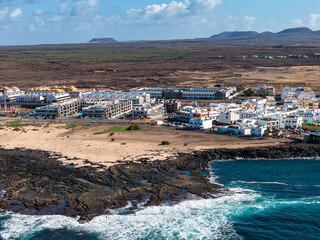 Aerial view of Fuerteventura shows whitewashed buildings and modern resorts by a rocky lava shore and sandy beach, turquoise surf, volcanic cones, and clear midday skies. © Aerial Film Studio