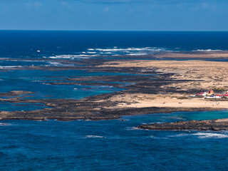 Aerial view shows Fuerteventura north coast, Playa del Bajo de la Burra, turquoise lagoons, volcanic reefs, whitewashed buildings, choppy Atlantic swells at midday. © Aerial Film Studio