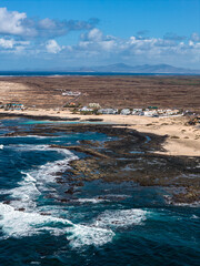 Aerial view shows turquoise Atlantic surf on dark lava reefs and pools, whitewashed houses by pale sand, arid plain, distant mountains, and hazy Lanzarote. © Aerial Film Studio