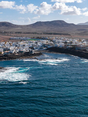 Aerial view shows whitewashed town on dark lava shore in Fuerteventura, Atlantic waves churn, black rock coves and turquoise surf, midday sun, scattered cumulus clouds. © Aerial Film Studio