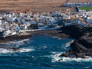 Aerial town of white buildings lines a rocky bay in Fuerteventura. Volcanic cliffs, crashing waves, and a small cove sit by a sports field under clear daylight. © Aerial Film Studio