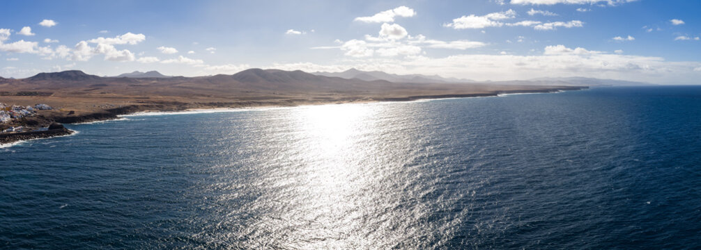 Aerial view shows Fuerteventura northwest coast at midday, a whitewashed village on a rocky headland, long beaches, cliffy points, and caldera silhouettes.