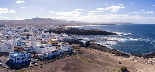 Fototapeta premium Aerial view shows white flat roof houses on black volcanic rock by the Atlantic, rugged lava reefs, turquoise surf, sandy beaches, and arid mountains in Fuerteventura.