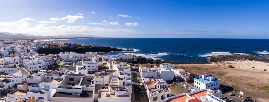 Aerial view shows whitewashed cubic houses by the Atlantic on Fuerteventura, Canary Islands, with black volcanic rock shelves, sandy plain, and midday clear skies.