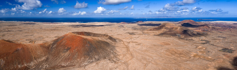 Aerial view of Fuerteventura badlands, a rust cinder cone crater, rounded cones to the Atlantic, northern plains toward Lanzarote, dunes, farms, midday light, cumulus clouds.