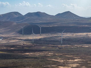 A wide aerial view shows tall wind turbines on arid plains in Fuerteventura, Canary Islands, with rounded cinder cones, dark lava fields, and soft afternoon light.