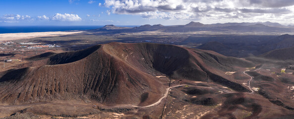 Aerial view of a dark horseshoe caldera, dunes, whitewashed towns, and wind turbines on Fuerteventura, facing the north coast and Atlantic under cumulus clouds.