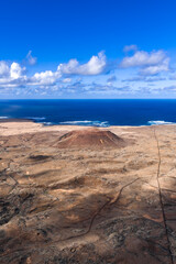 Aerial view shows a solitary cinder cone on Fuerteventura, near Caldera Los Arrabales, with tracks and fissures, low clouds, white surf, and bright midday light.