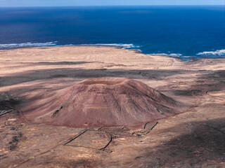 Aerial view shows a solitary cinder cone on a rust colored lava plain, broad summit crater, smooth flanks, pale coastal dunes, and the Atlantic in harsh midday light.