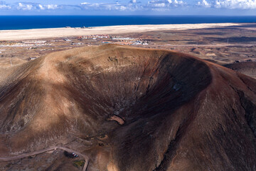 Aerial view of a volcanic crater with rust walls and pale floor, Fuerteventura, Canary Islands, Atlantic and sand dunes, midground settlement, daylight and clouds.