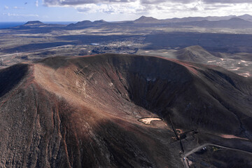Aerial crater view in Fuerteventura shows caldera, erosion lines, pale mineral patch, access road, cars, volcanic cones, Atlantic coast, town, wind turbines, late day light.
