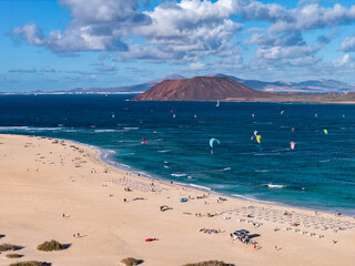 Crisp daylight aerial view shows golden dunes, a wide sandy beach, kitesurf sails, sunbathers, and Isla de Lobos with Lanzarote on the horizon in the Canary Islands.