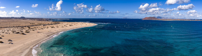 Aerial view shows Corralejo dunes, turquoise shallows, Isla de Lobos, and Lanzarote. Kites trace arcs over waves in late day light with scattered clouds above.