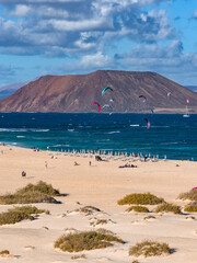 Kite surfers skim turquoise waters by Corralejo dunes, Fuerteventura. Colorful kites arc as sunbathers and umbrellas line the sand, with Isla de Lobos and a sailboat present.
