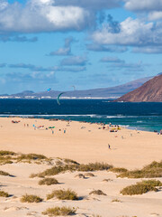 Aerial view of Fuerteventura, Canary Islands, with kite surfers, dunes, sparse shrubs, Strait of La Bocaina, Lanzarote silhouette, and a volcanic hillside in crisp daylight.