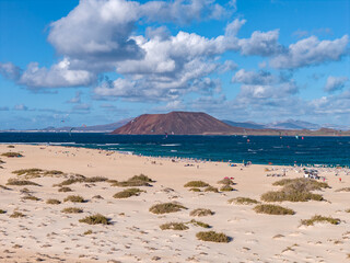Wide sandy beach with shrubs meets a deep blue strait on Fuerteventura. Kitesurf sails arc offshore as beachgoers walk dunes, with Isla de Lobos and Lanzarote.