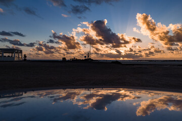 Golden clouds radiate crepuscular rays over the Atlantic at Fuerteventura. Silhouettes of a pavilion, low structures, and flagpoles reflect on a glossy surface.