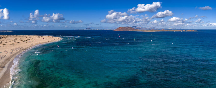 Aerial view shows Corralejo dunes curving to turquoise water, Isla de Lobos cone, and Lanzarote horizon. Kite sails streak over Playa del Bajo de la Burra at midday.