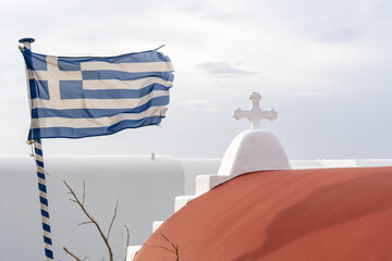 View of a Greek flag waving proudly against the whitewashed buildings with a red dome and cross, contrasting with the bright sky, Mykonos, Greece.
