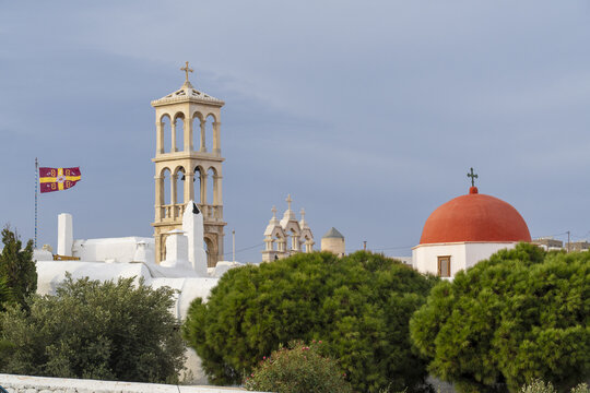 View of the iconic red dome and bell tower of monastery rise amidst the whitewashed buildings and lush greenery under a serene sky, Ano Mera, Mykonos, Greece.
