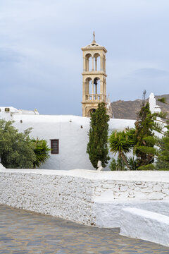 View of stark white buildings contrast with the earthy tones of a bell tower of monastery and surrounding greenery under a soft, overcast sky, Ano Mera, Mykonos, Greece.
