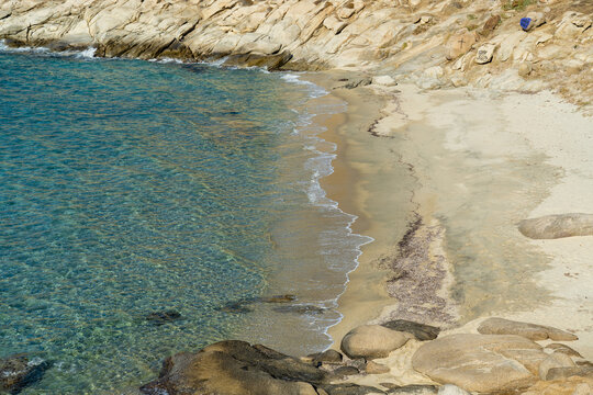 View of turquoise waters meeting the sandy shore, framed by rugged rocks and sun-kissed sands, a serene coastal scene unfolds in Mykonos, Greece.