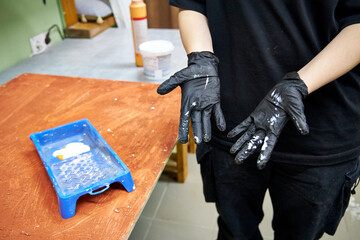 Close-up of a person wearing black gloves painting a wooden surface indoors. DIY home renovation process with brush and paint tray, workshop atmosphere, creative manual work and interior improvement c