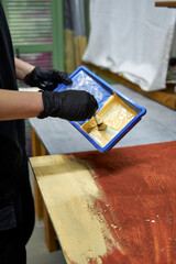 Close-up of a person wearing black gloves painting a wooden surface indoors. DIY home renovation process with brush and paint tray, workshop atmosphere, creative manual work and interior improvement c