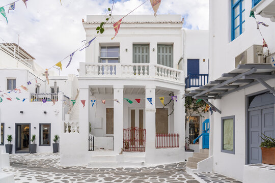 View of a charming, sun-drenched street lined with whitewashed buildings adorned with colorful flags contrasting against the patterned stone ground, Mykonos, Greece.