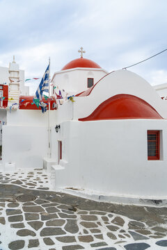 View of a Greek flag waving beside stark white buildings with red domed roofs under a cloudy sky along a stone tiled street, Old town, Mykonos, Greece.