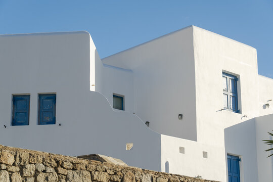 View of white buildings with blue window frames against a clear blue sky, above a stone wall in Old Town, Mykonos, Greece.