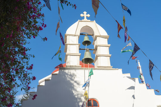 View of whitewashed church with a striking red dome, topped with a cross, framed by vibrant bougainvillea and festive flags, Mykonos, Old town, Greece.