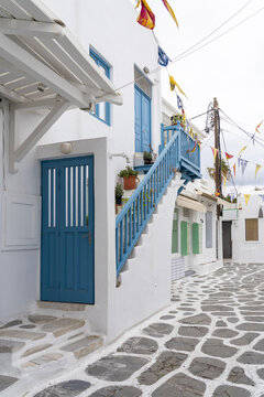 View of a bright blue door and matching staircase accenting the stark white buildings along a cobblestone street decorated with colorful flags, Old town, Mykonos, Greece.