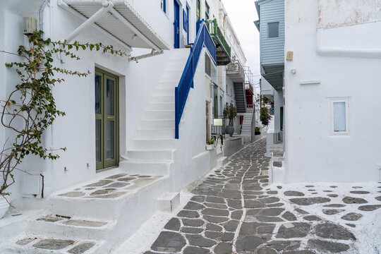 View of whitewashed buildings with vibrant blue and green accents line a narrow cobblestone street under a cloudy sky, Old town, Mykonos, Greece.