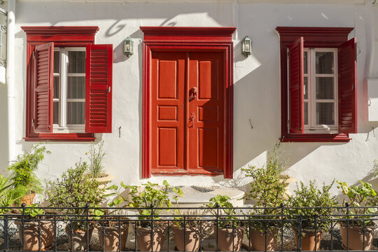View of the bright red door and window frames contrasting against the stark white walls, framed by potted plants creating a vibrant scene, Mykonos, Greece.