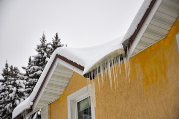 Icicles hanging from a snow-covered house roof in winter. Frozen ice formations on the eaves of a country home after snowfall, cold weather hazard, seasonal background with copy space