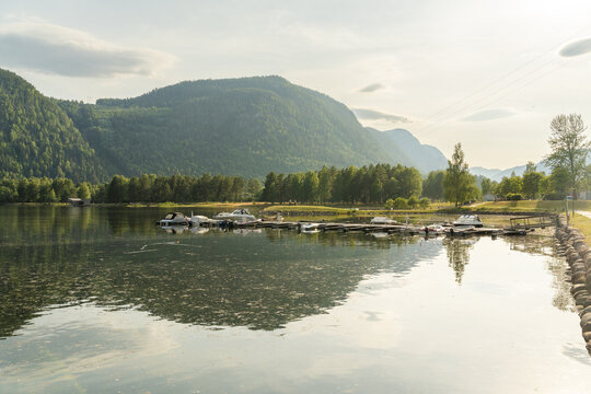 View of boats docked along a peaceful lake reflecting the verdant mountains and shoreline under a soft sky, Dalen, Telemark, Norway.