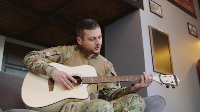 Soldier plays acoustic guitar in a cozy indoor setting while wearing military uniform during a break from duty