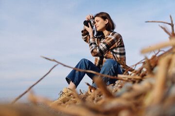 Woman photographer in a plaid shirt sits among dried reeds, aiming a camera toward the horizon in an outdoor nature setting with a calm blue sky and focused expression.