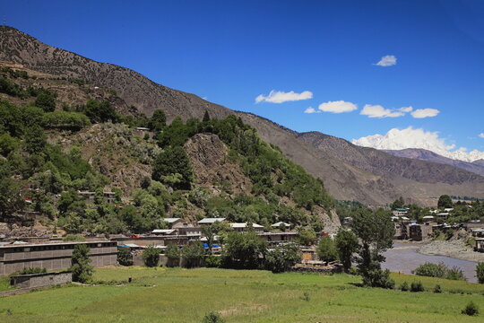 View of rugged, brown mountains cascade down to a valley dotted with small buildings, lush green fields, and a winding river under a brilliant blue sky, Chitral, Khyber Pakhtunkhwa, Pakistan.