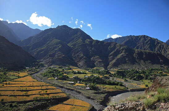 View of golden terraces meet the rugged, shadowed mountains under a vast blue sky, nestled in a valley of green and brown, Chitral, Khyber Pakhtunkhwa, Pakistan.