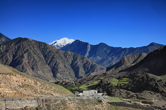 View of rugged, textured mountains under a brilliant blue sky, with a snowy peak in the distance casting shadows on the valley, Chitral, Khyber Pakhtunkhwa, Pakistan.