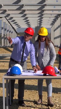 Construction Project Manager and Female Lead Architect Checking Blueprints at Construction Site. Man and woman in hardhats discuss a project at new industrial building construction site on a sunny day