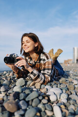 Woman photographer lying among smooth pebbles on a sunny beach with a camera, capturing a candid...
