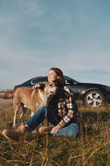 Fototapeta premium Woman sitting in a field with her dog, relaxing outdoors beside a car under a clear sky. Casual plaid shirt and jeans, a peaceful moment of companionship and nature on a roadside escape