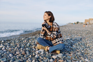 Woman sitting on a pebble beach with a camera, looking toward the sea, wearing a plaid shirt and jeans, capturing candid coastal moments and enjoying peaceful outdoor photography.